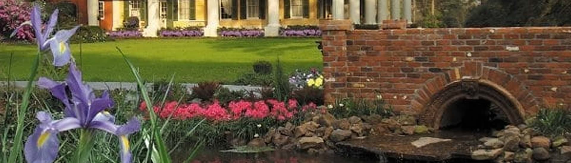 A picturesque garden with colorful flowers and a brick bridge leading to a serene pond, framed by a stately house in the background.