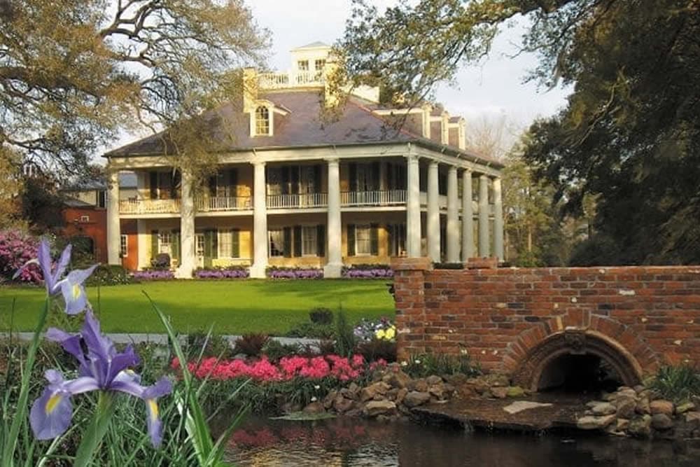 A grand, two-story mansion with a large porch, surrounded by colorful flowers and a brick bridge over a small water feature.