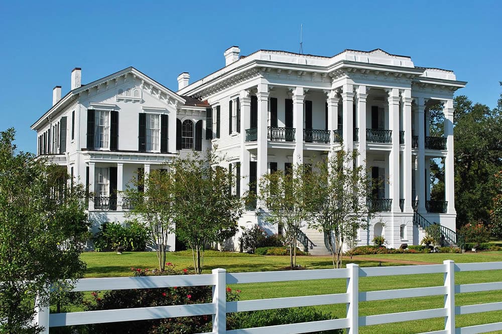 A grand white mansion with large columns, surrounded by greenery and a white fence.