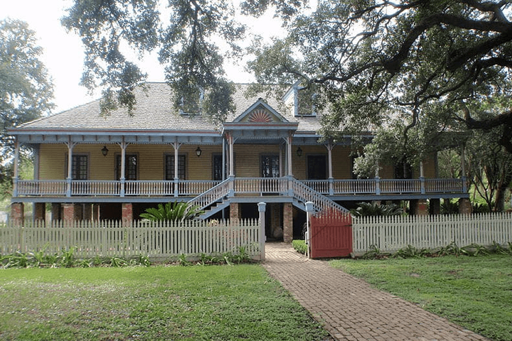 A historic yellow house with a wide front porch, set behind a white picket fence and surrounded by greenery.