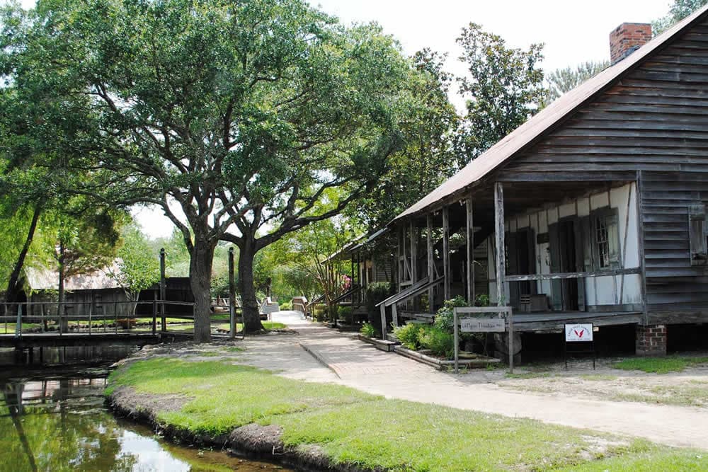 A historic wooden house beside a pathway and a tree, with a canal nearby.
