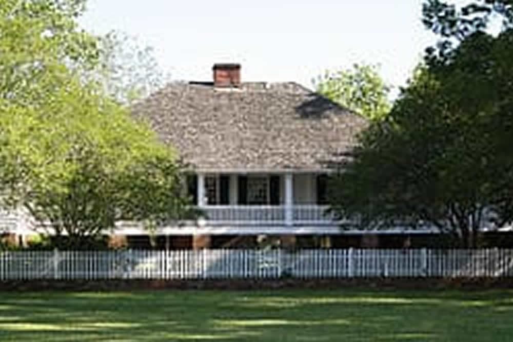 A large, two-story house with a pitched roof, surrounded by green trees and a white picket fence.