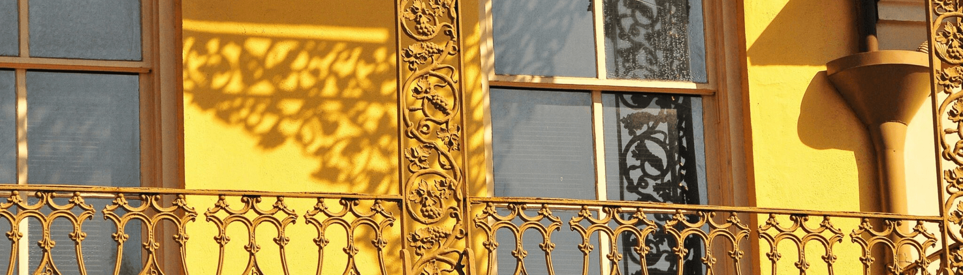 A vibrant yellow wall with a decorative wrought-iron balcony and intricate shadows cast by nearby foliage.