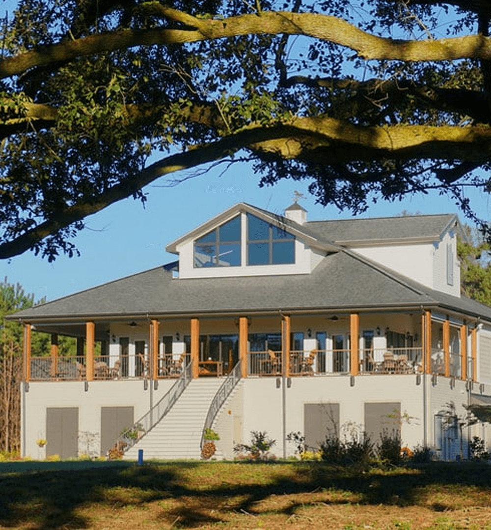 A modern two-story house with a wraparound porch, set against a clear blue sky and shaded by trees.