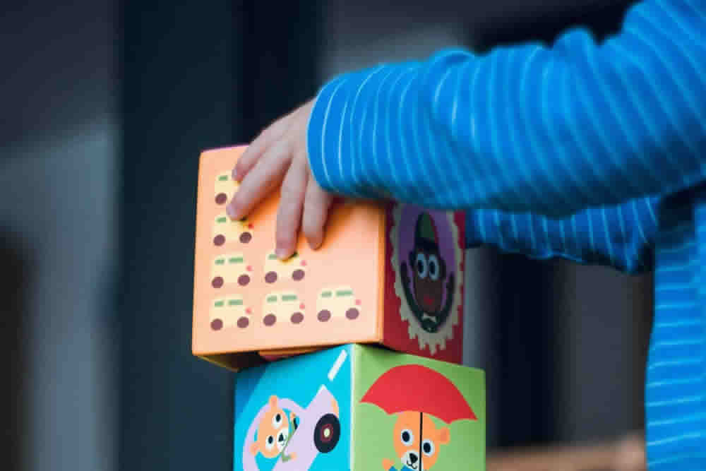 A child's hand places a brightly colored block on top of a stack of toy blocks.