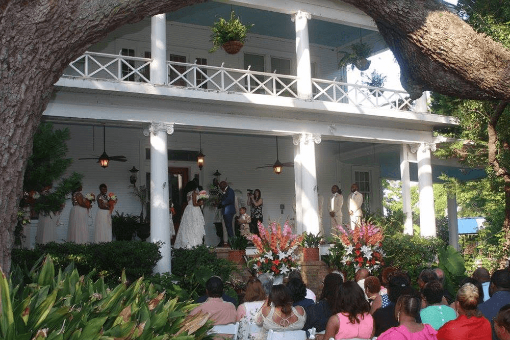 A wedding ceremony takes place on the porch of a charming white house adorned with floral decorations and surrounded by guests.