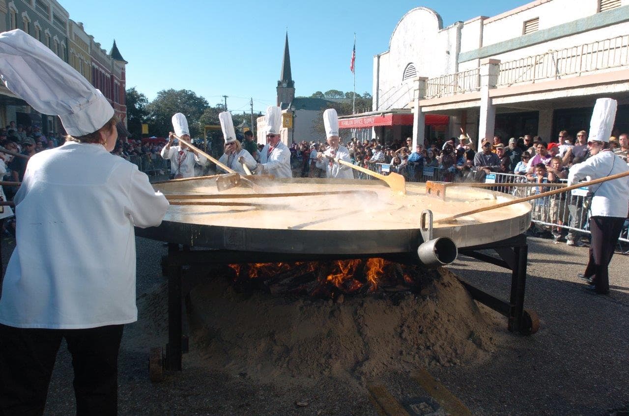 Chefs stir a large cauldron over an open flame at a crowded outdoor event.