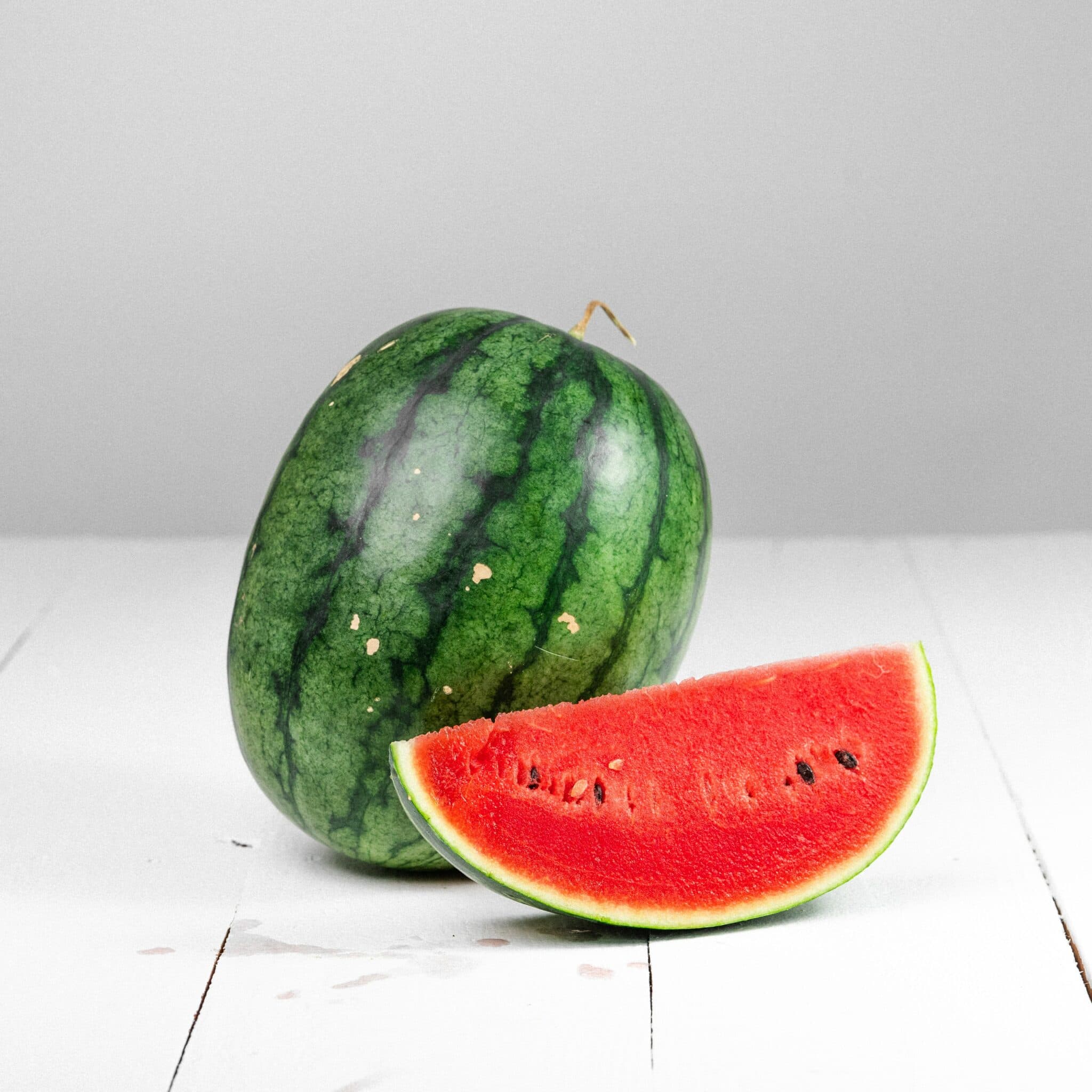 A whole watermelon and a slice of watermelon on a white wooden surface. A whole watermelon and a slice of watermelon on a white wooden surface.