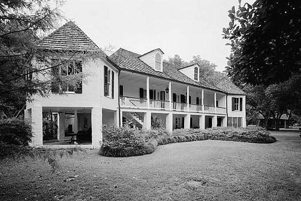 A historic two-story house with a porch surrounded by greenery, presented in black and white.