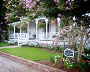 A charming white house with a porch, surrounded by blooming flowers and greenery.