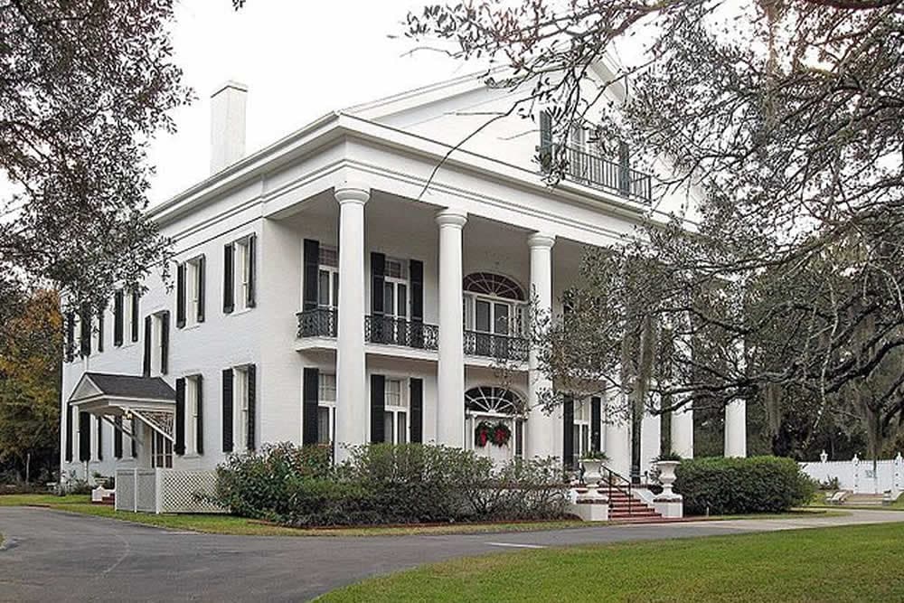 A large white colonial-style house with columns, surrounded by greenery.