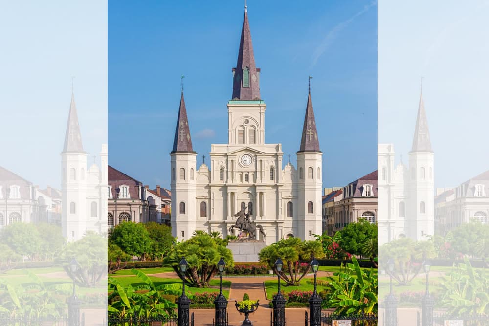 Historic building with towers and a clock, surrounded by greenery and statues.