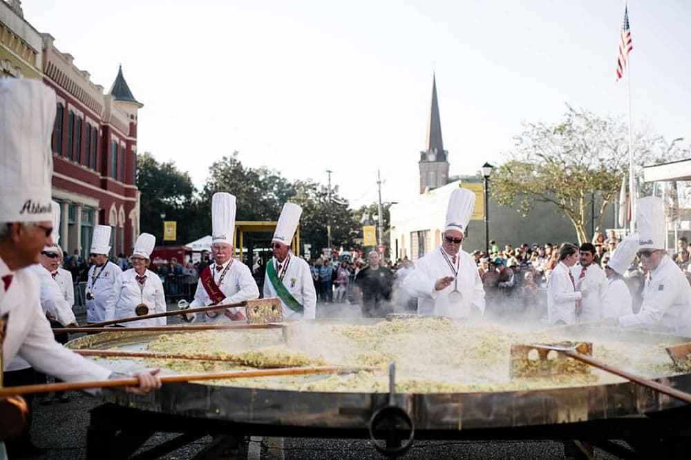 Chefs prepare a large pot of food at a bustling street festival.