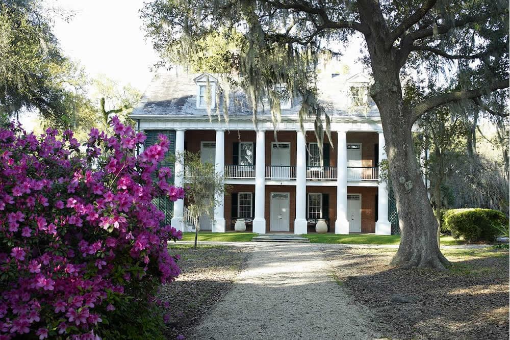 A historic two-story house with white columns, surrounded by vibrant pink flowers and lush greenery.