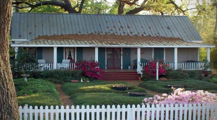 A charming, weathered house with a metal roof surrounded by lush gardens and a white picket fence.
