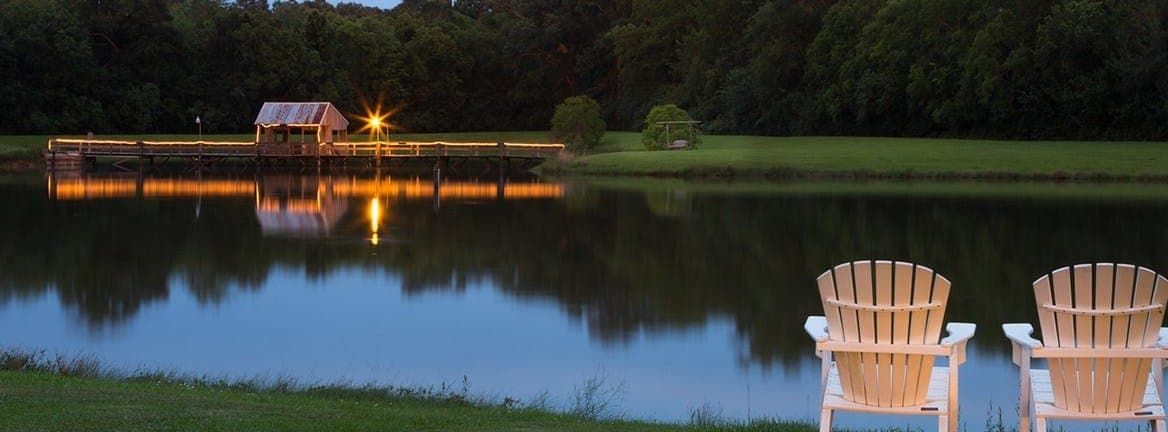 Two white Adirondack chairs face a peaceful lake, reflecting a nearby cabin and surrounding greenery at dusk.