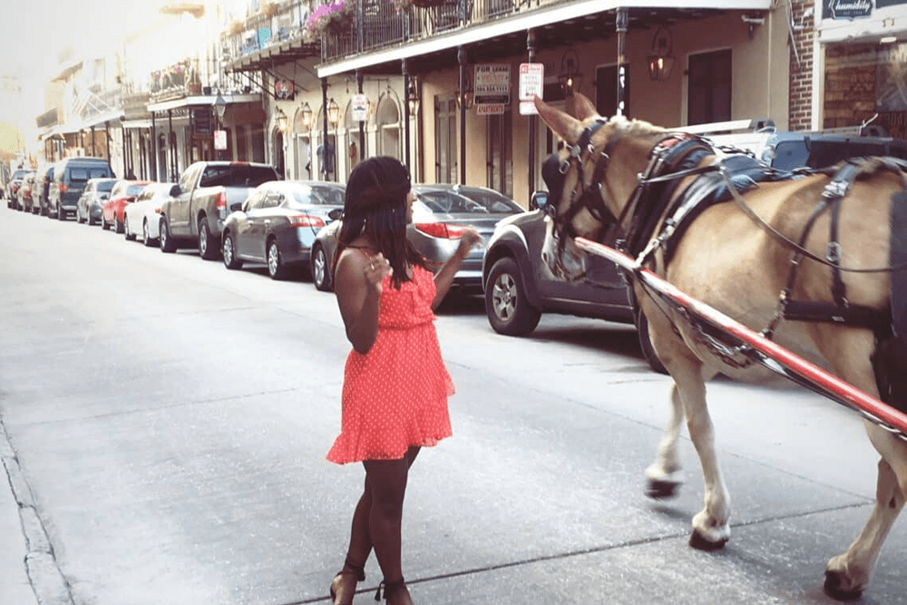 A girl in a red polka dot dress stands on the street as a horse-drawn carriage passes by.