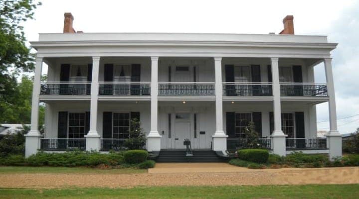 A large, white, two-story historic house with a porch supported by columns and manicured landscaping.