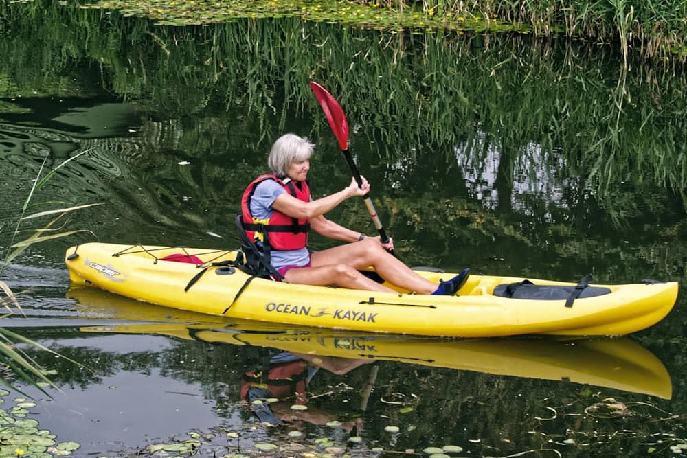 A woman paddles a yellow kayak on a calm, reflective water surrounded by greenery.
