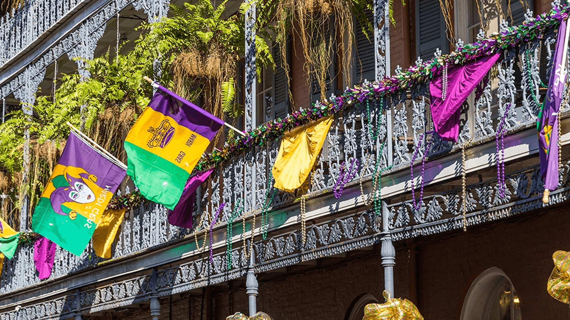 Colorful Mardi Gras flags and decorations hang from a wrought-iron balcony adorned with greenery.