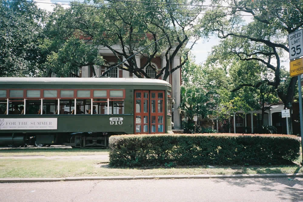 A vintage green streetcar with red windows stops by lush greenery and a historic building.