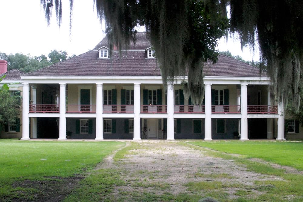 A stately plantation house with columns and a sloping roof, surrounded by greenery.