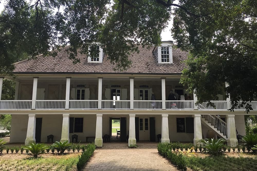 Historic two-story house with a porch, surrounded by trees and greenery.