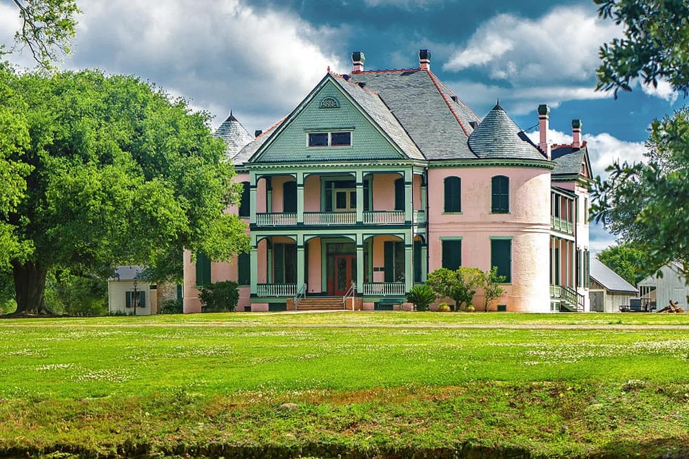 A large, pastel-colored Victorian mansion surrounded by lush greenery.